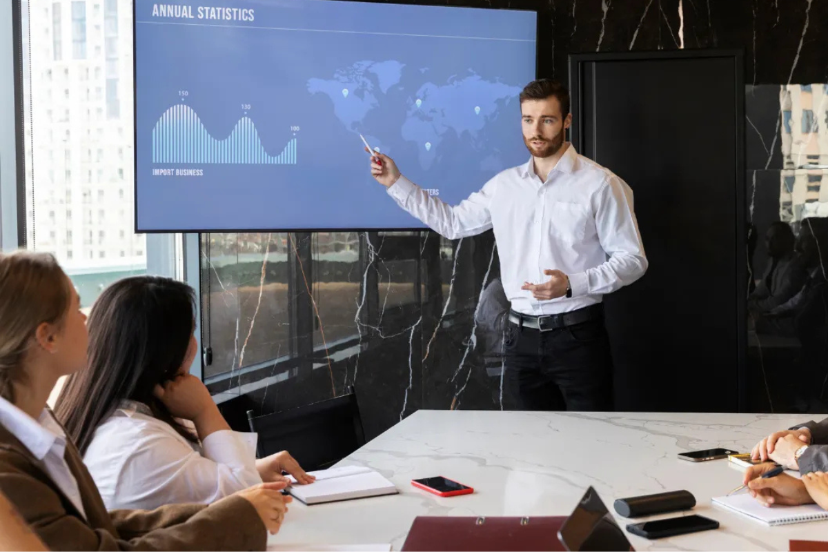 A man in a white shirt presenting annual statistics on a screen in a modern conference room, pointing to a graph and world map. Three colleagues seated at a table listen and take notes.